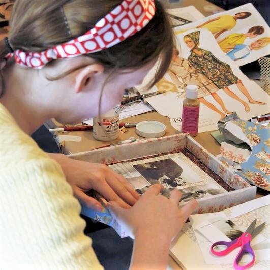 A person wearing a headband and pale yellow sweater is working on a mixed-media assemblage work. 